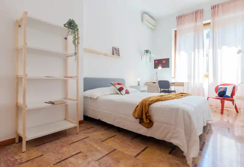 Dormitorio luminoso y bien decorado con cama doble, ropa de cama clara, un pequeño escritorio junto a la ventana y detalles cálidos. La ventana amplia con cortinas traslúcidas inunda la habitación de luz natural, perfecta para un anuncio de alquiler.
