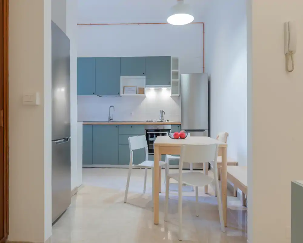 Modern compact kitchen with muted teal cabinets, stainless steel appliances and a small wooden dining table with chairs — bright, well-lit space ideal for a small household.