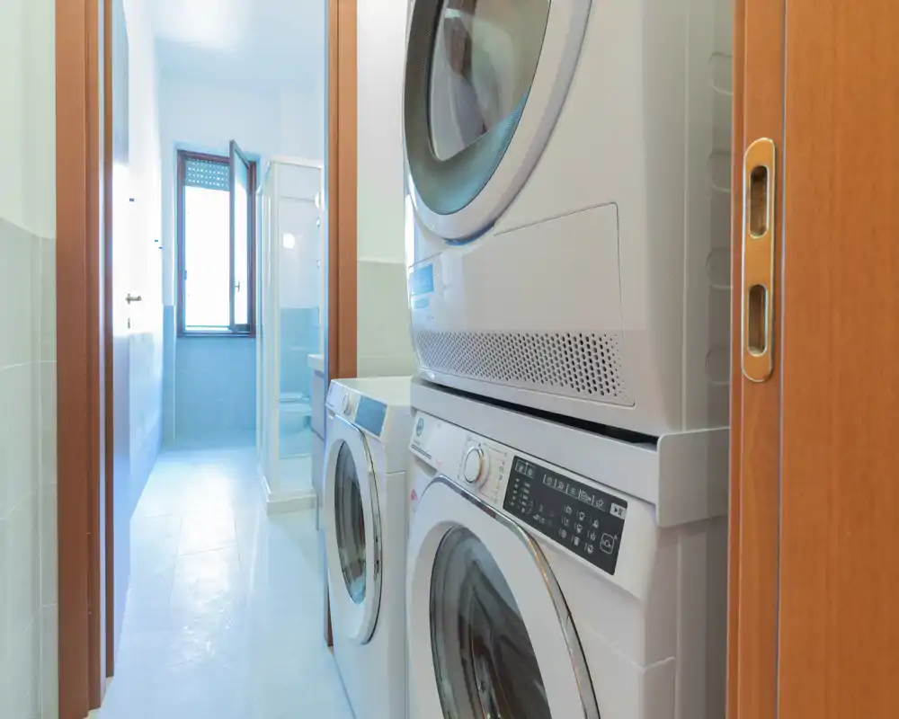A narrow bathroom with a stacked washer and dryer positioned near the doorway; a shower enclosure and window are visible at the far end.