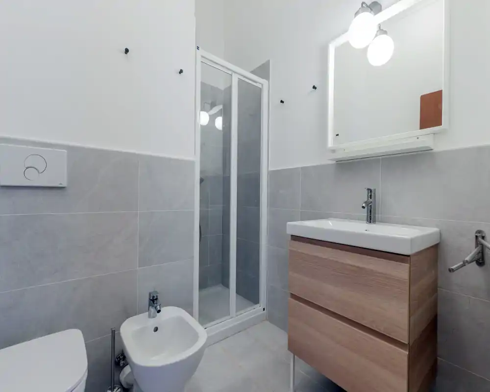 Modern compact bathroom with a shower cabin, bidet, toilet and a wooden vanity with sink; light grey tiles and a lit mirror create a clean, contemporary look.