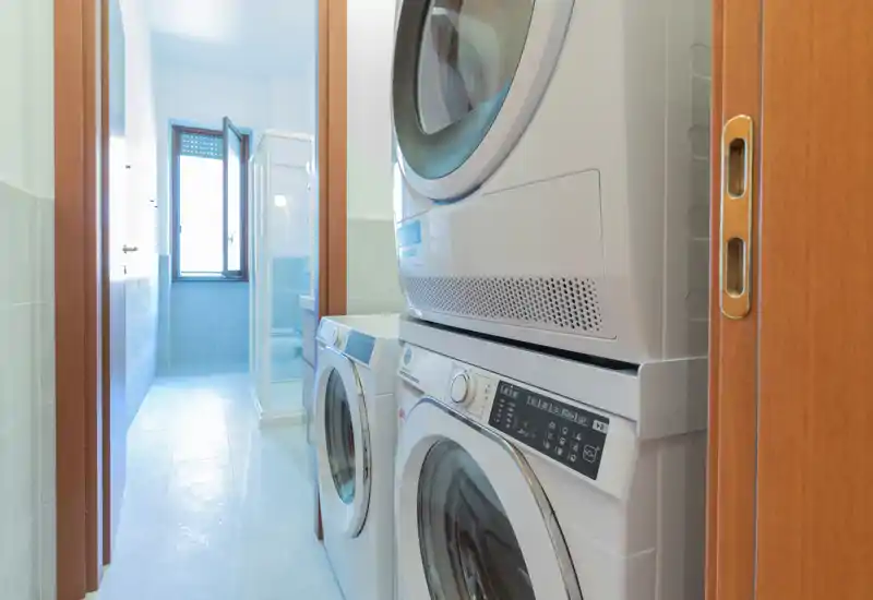 Bagno stretto con lavatrice e asciugatrice impilate vicino alla porta; in fondo si vede la cabina doccia e una finestra.