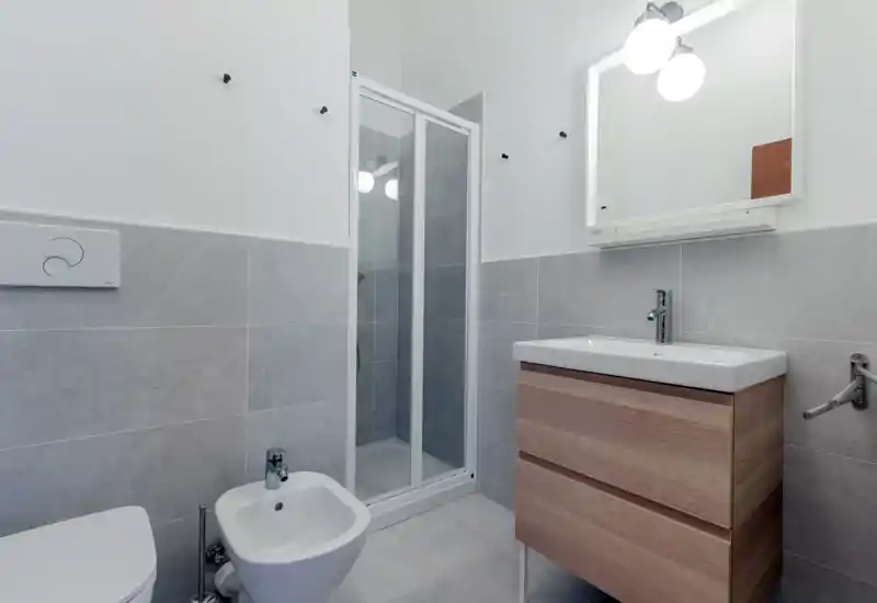 Bagno moderno e compatto con cabina doccia, bidet, WC e mobile lavabo in legno; piastrelle grigio chiaro e specchio con luce per un aspetto pulito e contemporaneo.