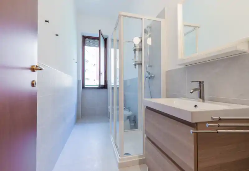 Bagno moderno e luminoso con box doccia angolare, mobile lavabo sospeso e finestra che garantisce luce naturale.