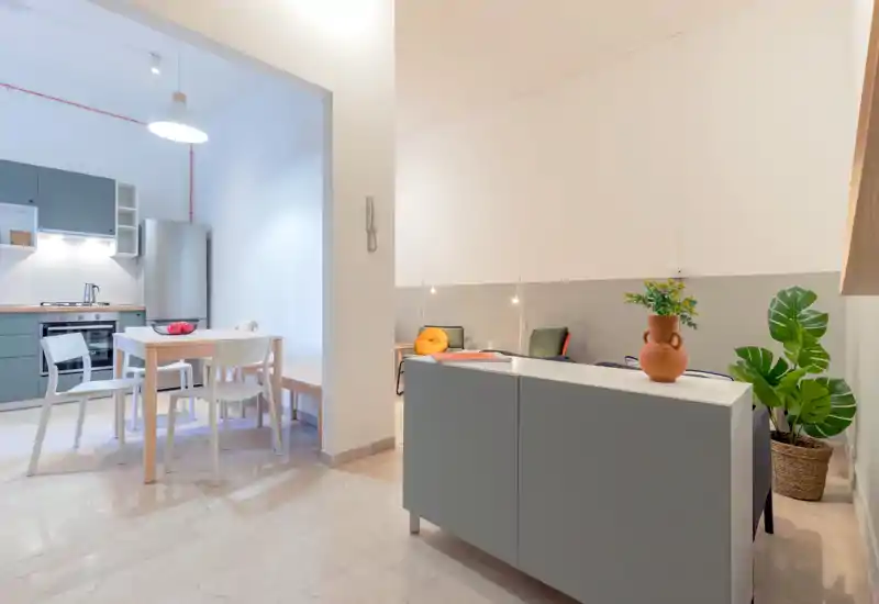Zona giorno moderna open-space luminosa con tavolo da pranzo e angolo cottura visibile. Arredi minimal in stile scandinavo, piante in vaso e dettagli morbidi rendono l'ambiente accogliente.