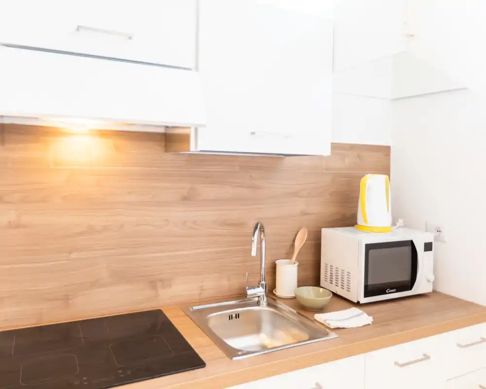 Angolo cottura luminoso e moderno con paraschizzi effetto legno, piano a induzione, lavello, microonde e bollitore — spazio minimale e ben arredato ideale per soggiorni brevi.