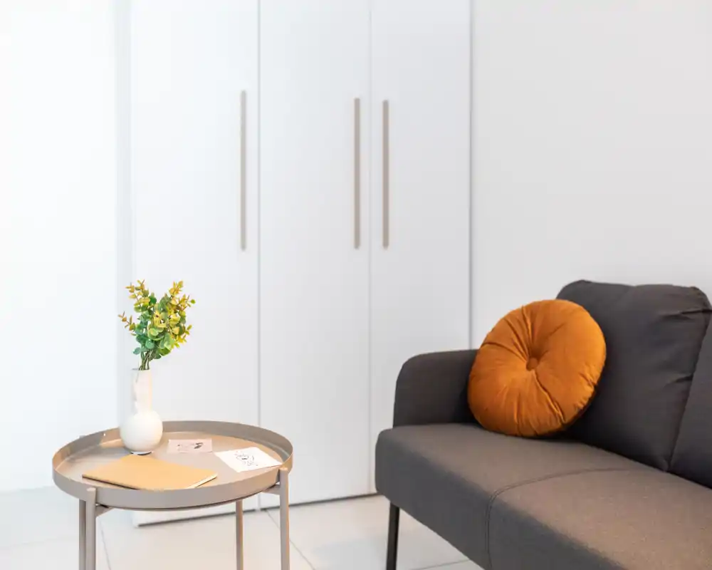 Zona giorno luminosa e minimale con un divano grigio moderno valorizzato da un cuscino rotondo caldo, un tavolino laterale e un vaso con fiori: stile pulito e accogliente, perfetto per un annuncio.