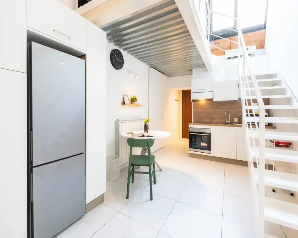 Cucina moderna e compatta con forno integrato, frigorifero e un piccolo tavolo da pranzo sotto il soppalco; design luminoso e pulito ideale per un monolocale.