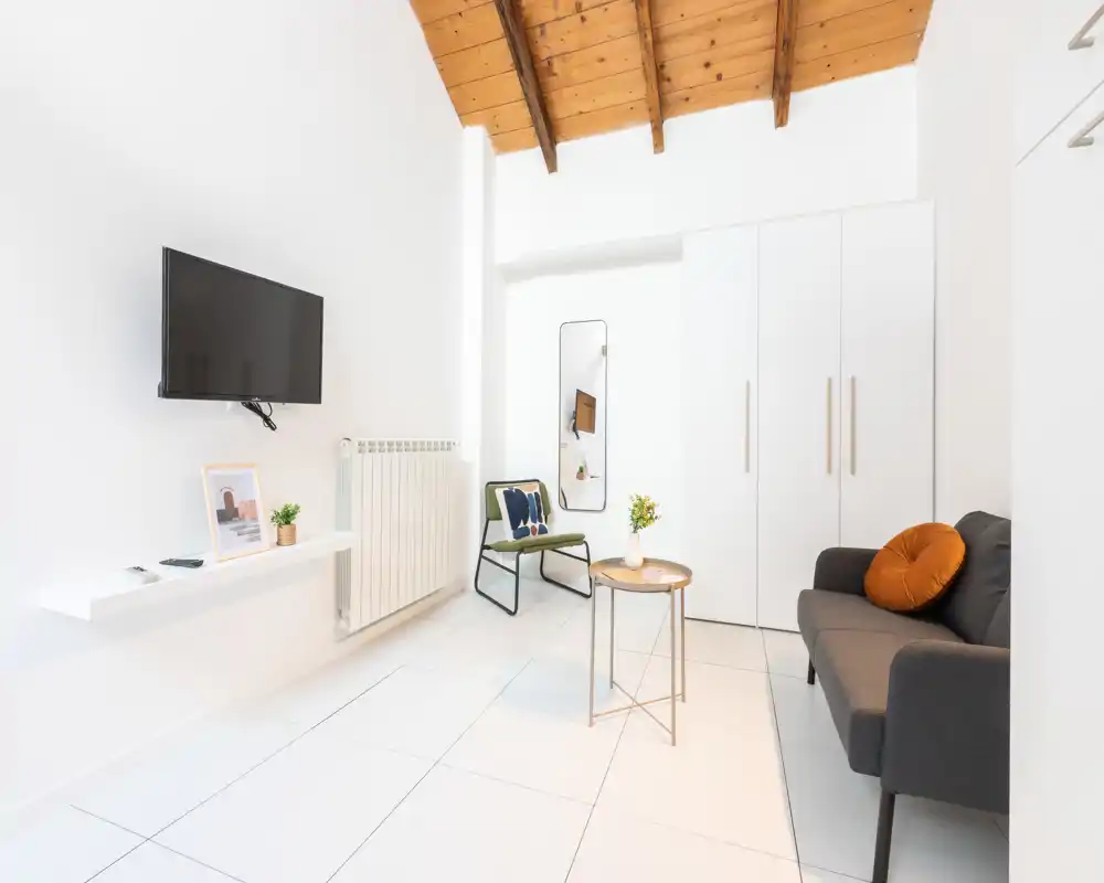 Area giorno luminosa e minimal con soffitto con travi in legno, divano compatto con cuscino decorativo, piccolo tavolino e TV a parete — perfetta per un affitto breve di stile.