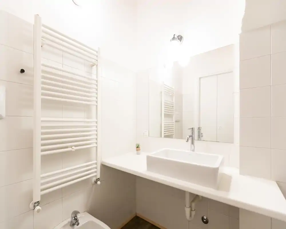 Bagno pulito e moderno con lavabo a bacinella bianco, grande specchio e termoarredo a soffietto; ambiente luminoso e minimal ideale per annunci.