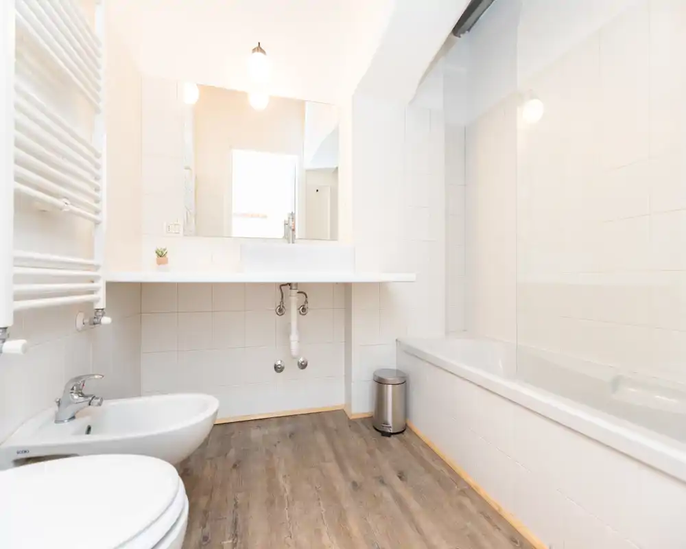 Bagno moderno e luminoso con pareti piastrellate bianche, vasca con pannello in vetro, lavabo sospeso e bidet, pavimento effetto legno e grande specchio che amplifica la luce.