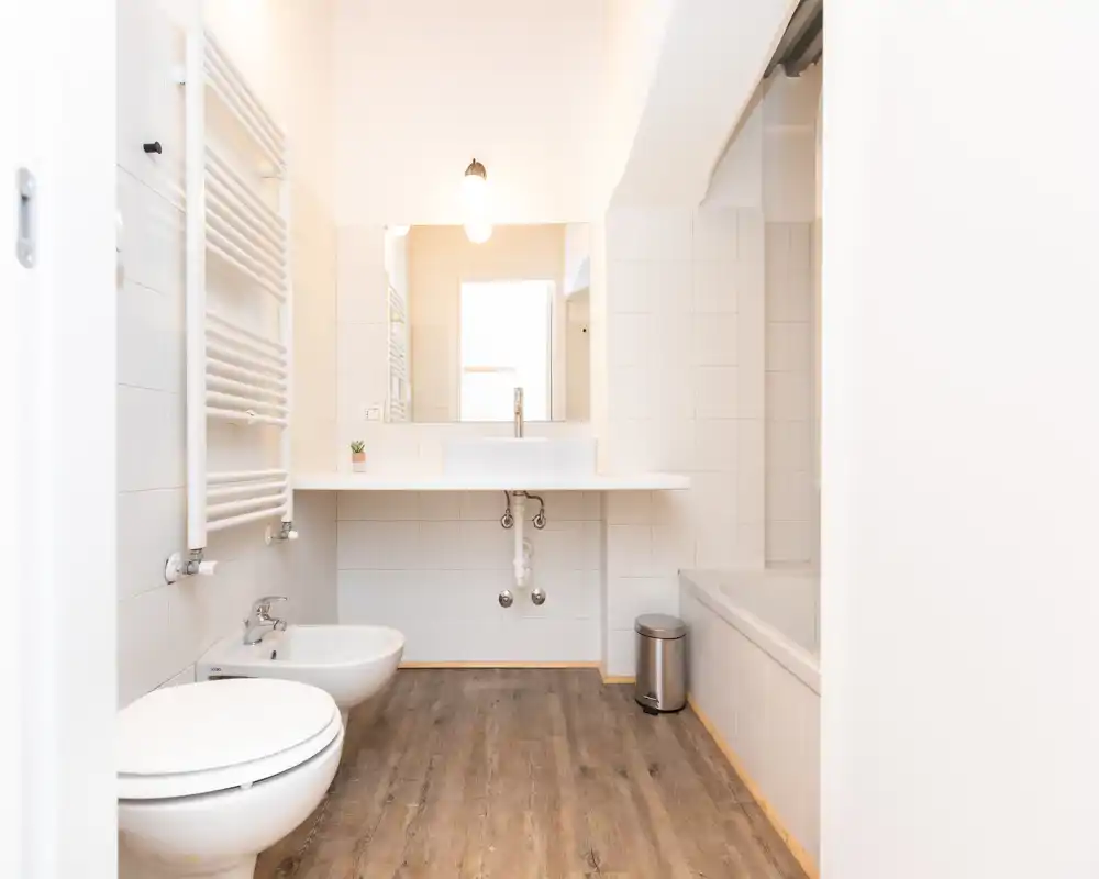 Bagno moderno e luminoso con WC, bidet, lavabo e vasca. Piastrelle bianche e pavimento effetto legno per un aspetto pulito e minimale.