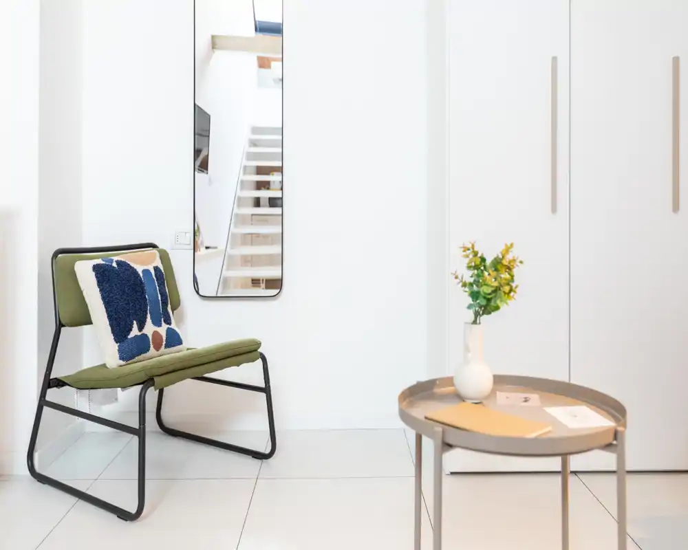 Ingresso minimal ben illuminato con una sedia d'accento, specchio a parete e un piccolo tavolino con vaso.