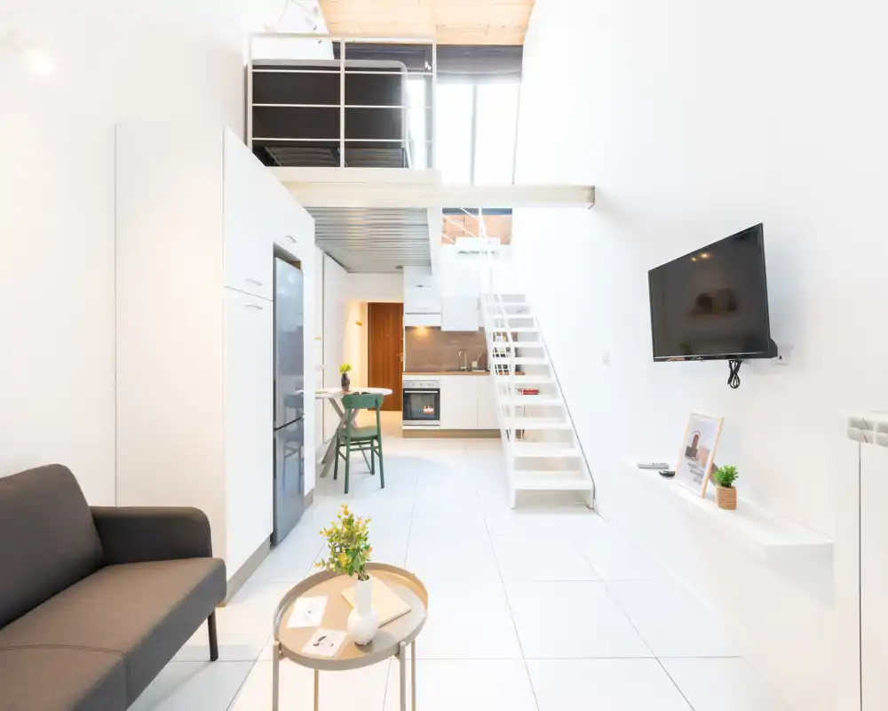 Zona giorno luminosa e moderna con angolo cottura compatto, pareti bianche e soppalco. L'ambiente presenta un divano scuro, TV a parete e pochi elementi decorativi per un look pulito e contemporaneo.