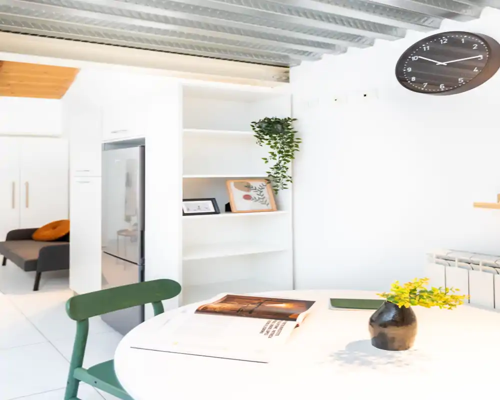 Angolo pranzo moderno e luminoso con tavolo rotondo bianco, sedia verde e semplici elementi decorativi come un vaso e scaffalature sullo sfondo.