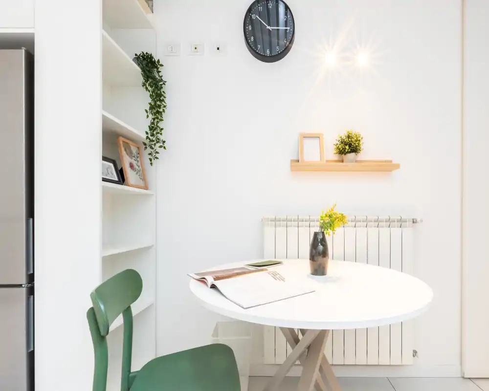 Angolo pranzo luminoso e minimale con tavolo rotondo bianco, sedie verdi e piccole piante decorative che creano un angolo fresco e accogliente ideale per pasti informali.