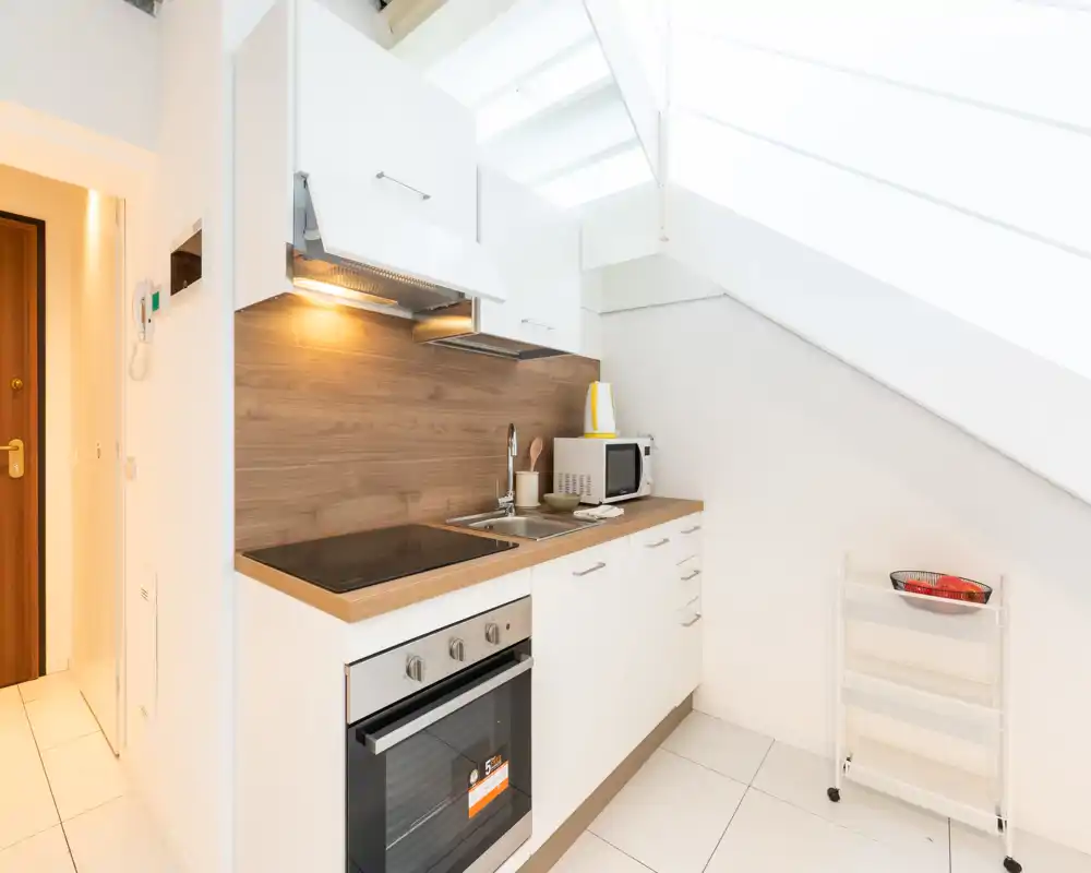 Angolo cottura moderno e compatto sotto una soffitta inclinata con mobili bianchi, schienale effetto legno, forno incorporato e piano a induzione. Layout luminoso, pulito e salvaspazio, ideale per un monolocale.