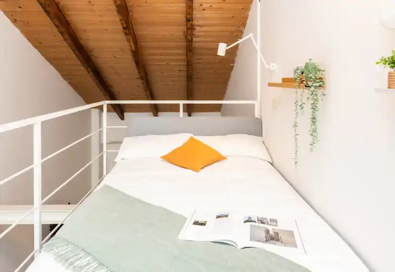Dormitorio en un loft luminoso y acogedor con cama bien hecha, vigas de madera cálidas y decoración moderna sencilla con cojín mostaza y plantas colgantes.
