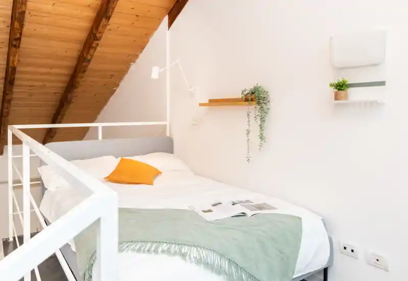Dormitorio tipo loft luminoso con cama doble cómoda, techo de madera inclinado y decoración moderna y sencilla — manta y cojín que crean un ambiente acogedor.