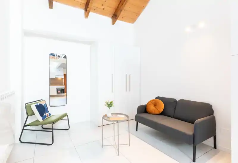 Zona de estar luminosa y minimalista con un sofá gris compacto, cojín decorativo y una butaca ligera. Paredes blancas y una pequeña mesa central crean una sensación moderna y aireada.