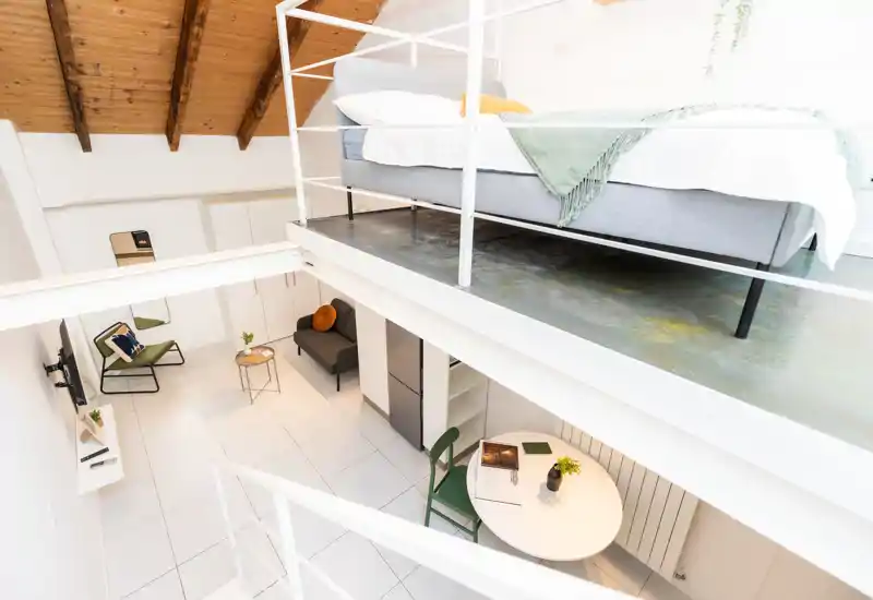 Dormitorio en loft luminoso con cama elevada en un altillo que domina la zona de estar; estilo moderno y minimalista, vigas de madera vistas y textiles acogedores.