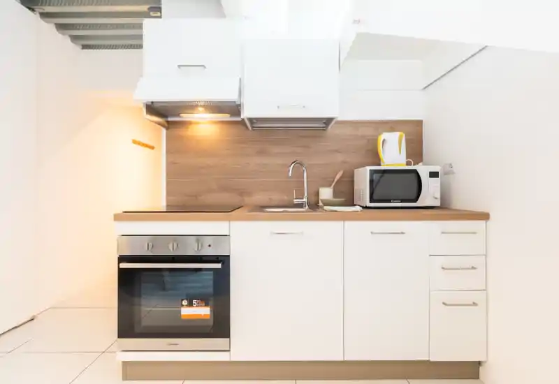Cocina pequeña y moderna con horno empotrado, placa de inducción, fregadero y microondas sobre encimera y salpicadero de madera — luminosa, limpia y minimalista.
