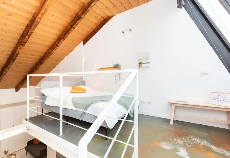 Dormitorio tipo loft luminoso con cama doble bajo vigas de madera cálidas, textiles suaves y una claraboya brillante — ideal para una estancia acogedora.