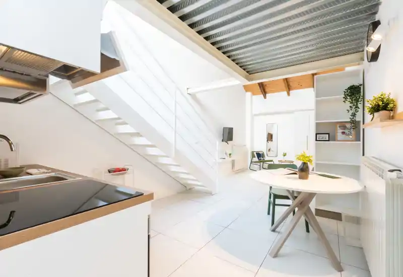 Cocina abierta moderna y luminosa con encimera compacta y fregadero integrado, junto a una pequeña mesa de comedor y escaleras — minimalista, elegante y bien iluminada.
