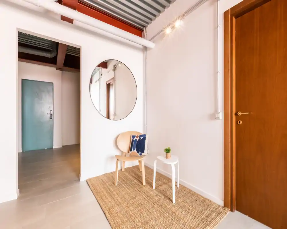 Ingresso accogliente con tappeto in fibra naturale, una sedia in legno con cuscino decorativo, uno specchio rotondo e una porta in legno caldo — ambiente luminoso e minimal.