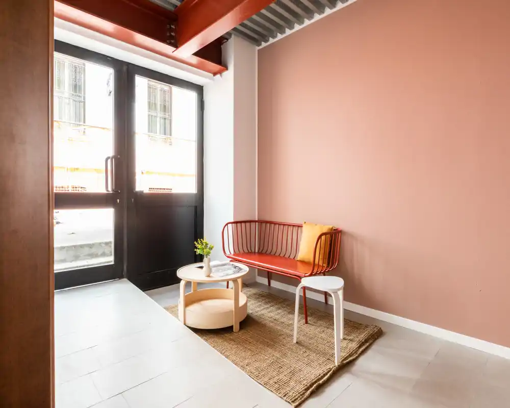 Ingresso moderno e luminoso con piccolo angolo seduta: panca rossa in metallo, tappeto in fibra, tavolino rotondo e sgabello accanto alla porta a vetri — atrio accogliente e dall'aspetto caldo.