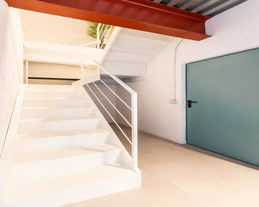 Ingresso luminoso e moderno con scale bianche pulite, corrimano in metallo e una porta turchese; spazio di passaggio minimal e ben illuminato, ideale per loft o studio.