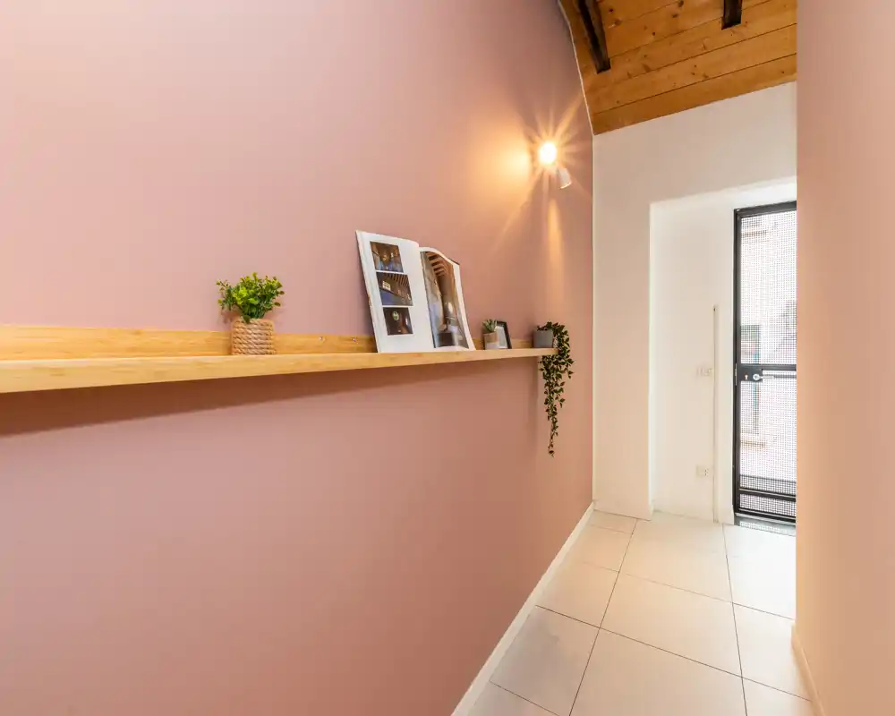 Ingresso luminoso e stretto con parete rosa calda, mensola in legno con piccole piante e oggetti decorativi, pavimento piastrellato e porta a vetro che lascia entrare luce naturale.