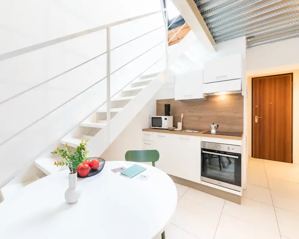 Angolo cottura moderno e compatto con ante bianche, forno integrato, microonde e un piccolo tavolo rotondo sotto una scala luminosa; design pulito e schienale in legno che rende lo spazio accogliente e funzionale.