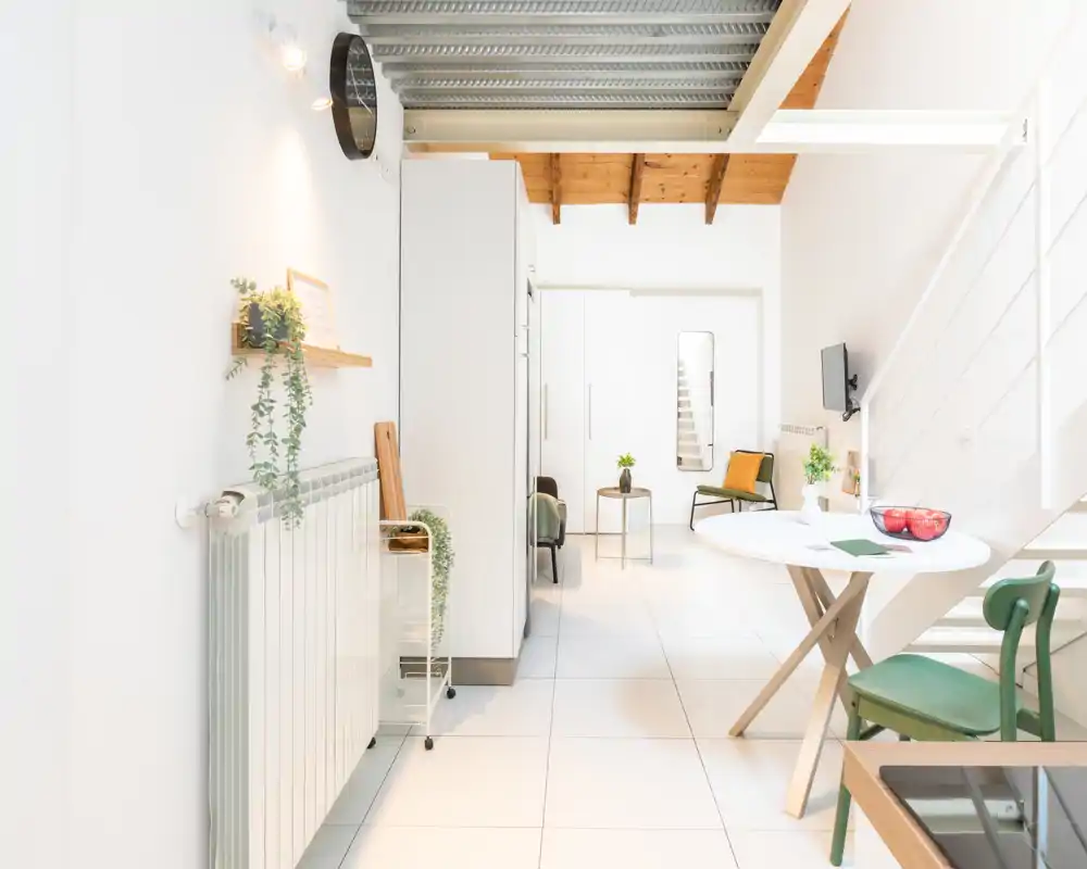 Zona giorno luminosa e moderna con pareti bianche, travi in legno, un tavolo rotondo e sedute confortevoli — ariosa e ben arredata per un affitto breve.