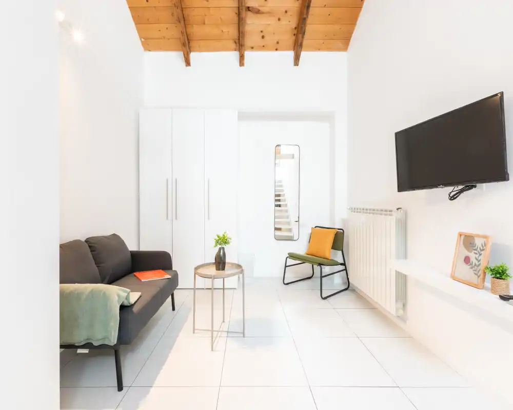 Zona giorno luminosa e moderna con un divano compatto, poltrona d'accento, TV a parete e travi in legno che donano un aspetto caldo e minimal.