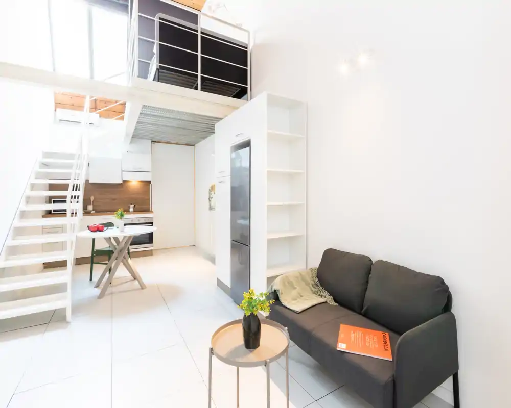 Zona giorno in stile loft moderna e luminosa con cucina compatta, scala verso il soppalco e divano grigio scuro; perfetta per affitti brevi in cerca di spazi open-plan contemporanei.