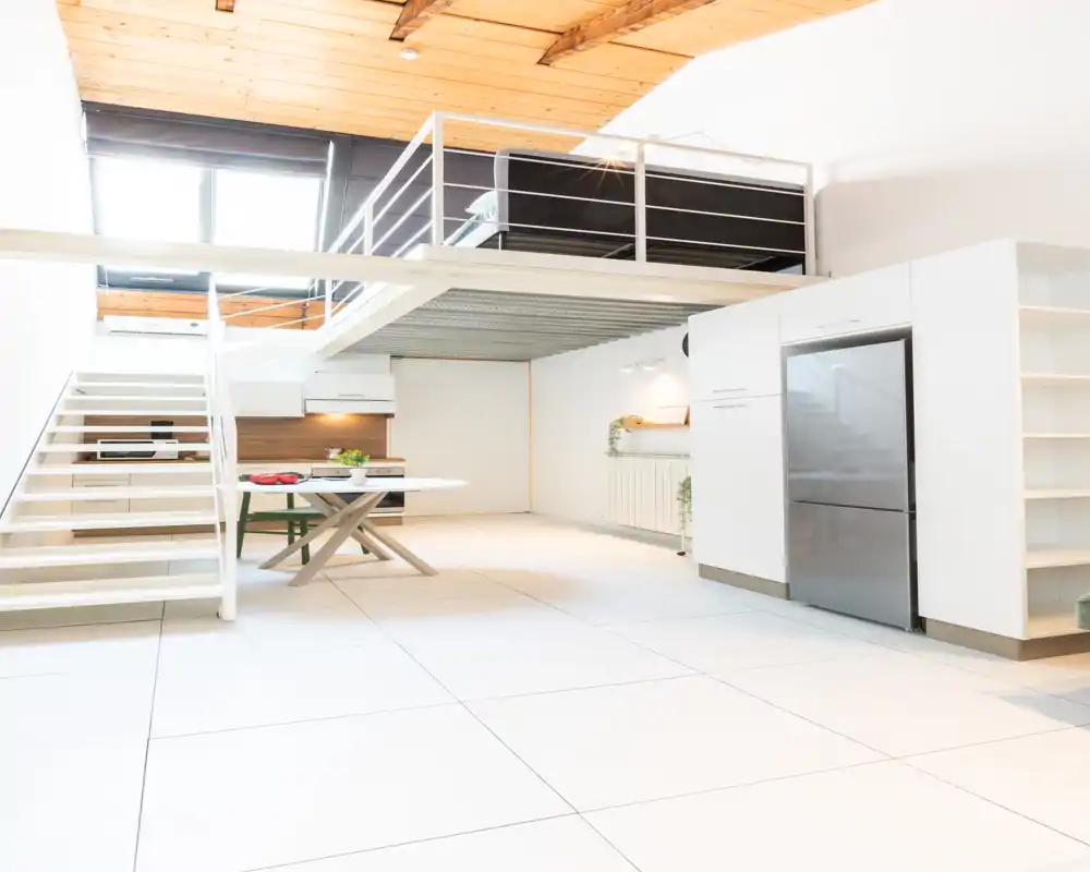 Ampio soggiorno moderno open-space con soppalco, tavolo minimal e elettrodomestici visibili; pavimento chiaro e soffitto in legno creano un'atmosfera luminosa da loft.
