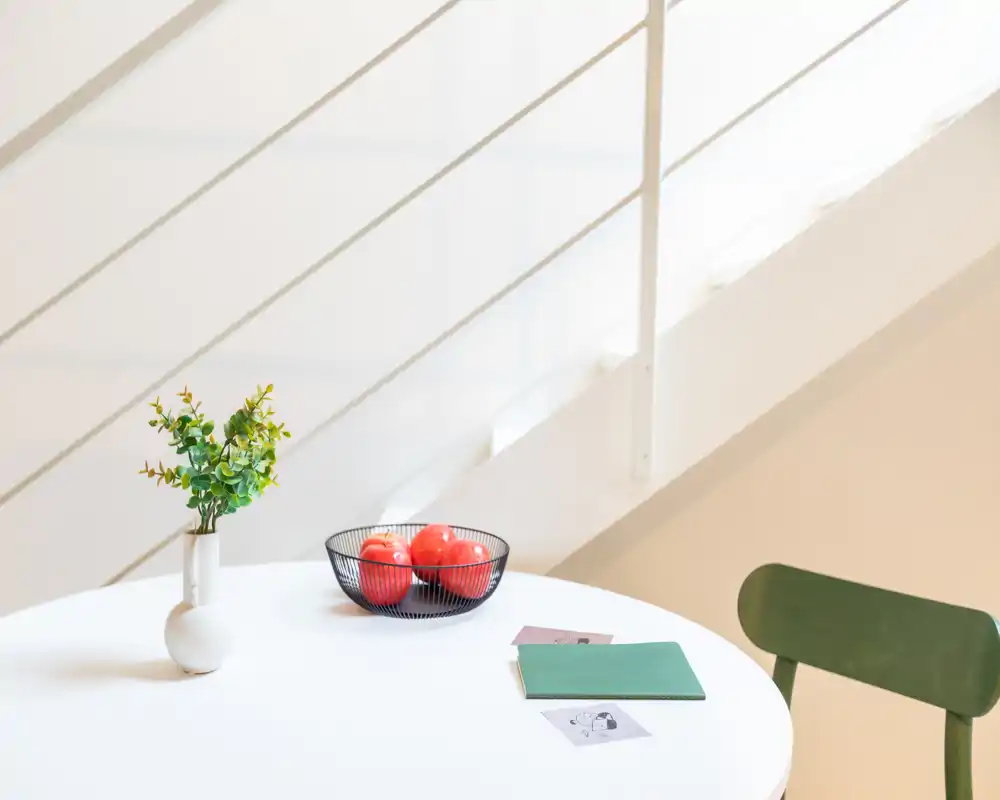 Angolo pranzo luminoso e minimal con tavolo bianco rotondo, vaso decorativo e ciotola di frutta — ambiente moderno e tranquillo perfetto per annunci.