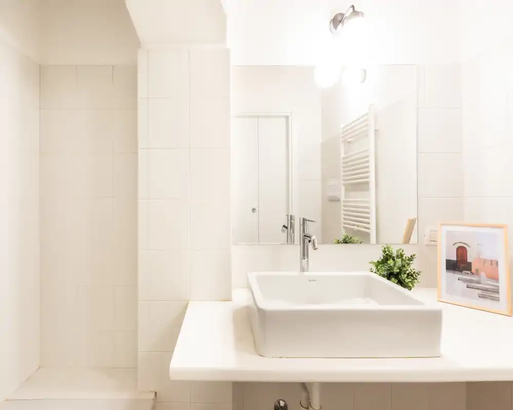 Zona lavabo bagno luminosa e moderna con lavabo rettangolare da appoggio, specchio a parete e pianta decorativa, in un ambiente piastrellato bianco e pulito.