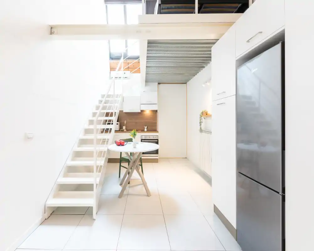 Cucina moderna e luminosa con un tavolo compatto, scale bianche che portano al soppalco e frigorifero in acciaio — pulita, ariosa e ideale per un piccolo appartamento.