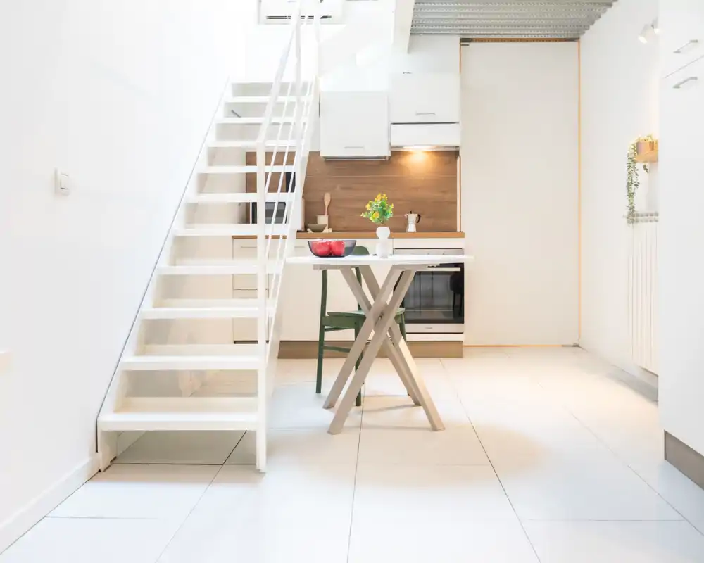 Cucina compatta e moderna con mobili bianchi, schienale in legno e un piccolo tavolo da pranzo sotto la scala — cucina da monolocale elegante e luminosa.