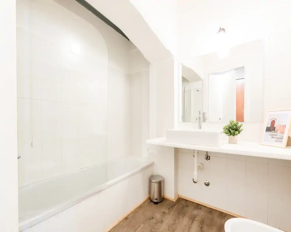 Bagno luminoso e moderno con vasca e schermo doccia in vetro, piastrelle bianche, lavabo a parete e pavimento effetto legno.