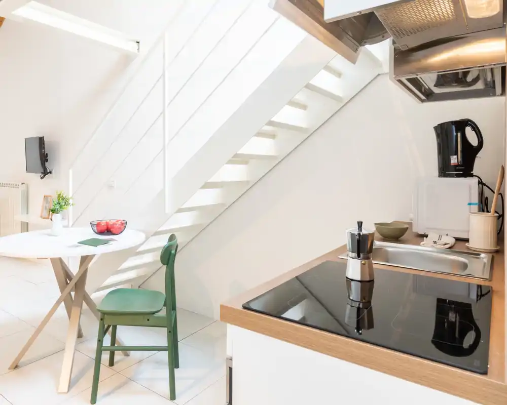 Angolo cottura moderno e compatto sotto la rampa di scale con piano a induzione, lavello e piccolo tavolo da pranzo. Stile luminoso e minimal con una sedia verde e dettagli decorativi.