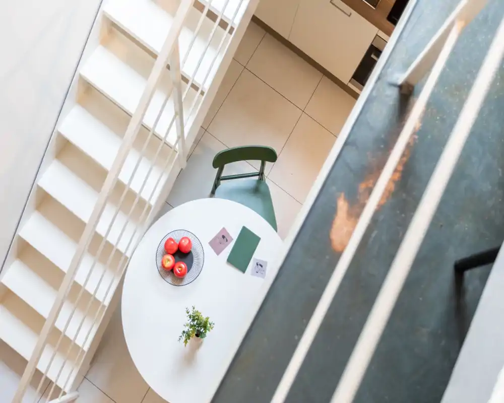 Vista dall'alto di un piccolo angolo cucina/pranzo con tavolo rotondo, sedia verde, ciotola di frutta e una pianta — minimal e ben illuminato.