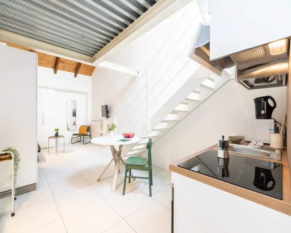 Zona giorno luminosa e moderna con angolo cottura compatto, tavolo da pranzo e scala — design minimale e arioso, perfetto per soggiorni brevi.