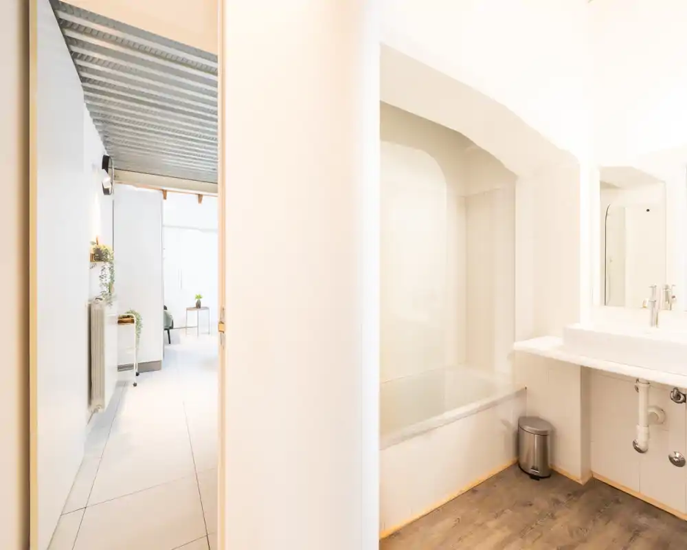 Bagno moderno e luminoso con vasca/doccia, lavabo bianco e specchio, pavimento effetto legno. Stile pulito e minimal con buona luce naturale, adatto per annunci di affitto.