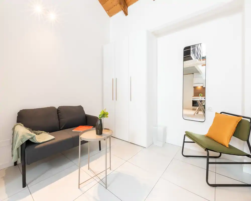 Zona giorno luminosa e moderna con un piccolo divano scuro, un tavolino rotondo e una poltrona accentata con cuscino giallo senape; pareti bianche e arredi minimal creano un ambiente ordinato e accogliente.