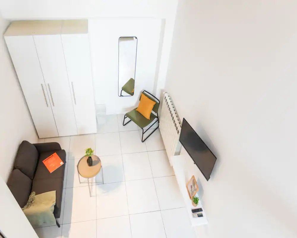 Zona giorno luminosa e moderna con divano compatto, piccolo tavolino e poltrona d'accento; mobili bianchi minimal e TV a parete creano un ambiente arioso e ben illuminato, ideale per un monolocale.
