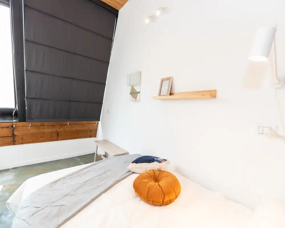 Camera da letto luminosa e minimalista con letto comodo, biancheria neutra, cuscini decorativi e dettagli in legno caldo vicino a una grande finestra con tenda scura.