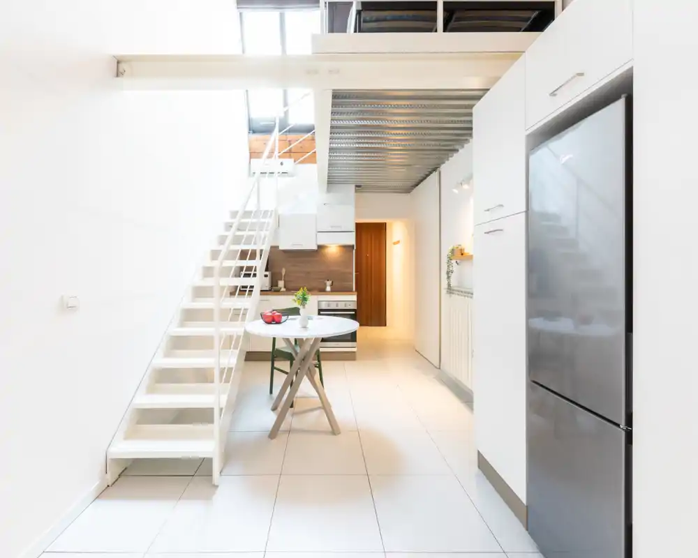 Cucina moderna open-space luminosa con piccolo tavolo da pranzo, frigorifero in acciaio e mobili bianchi; linee pulite e schienale in legno caldo rendono l'ambiente accogliente.