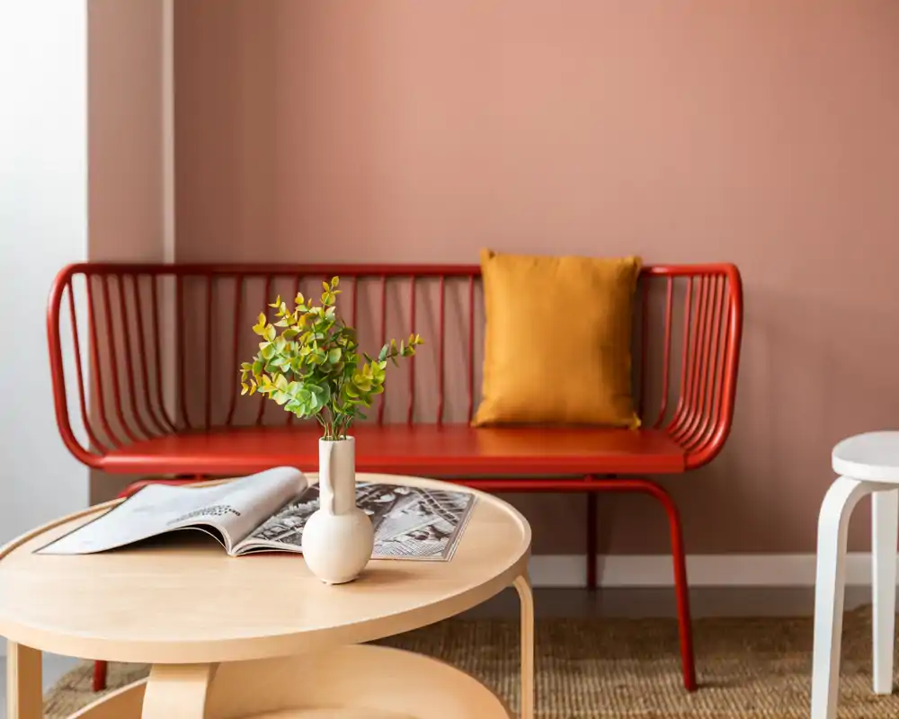 Piccolo soggiorno di stile con panca in metallo rossa, cuscino color senape e tavolino in legno chiaro con vaso decorativo e rivista aperta — ambiente caldo, luminoso e ben curato.